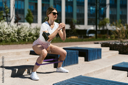 Young fitness woman in sportswear is training outdoors and doing a squat exercise with fitness elastic bands or an expander. Healthy lifestyle and sports  street training  social distancing