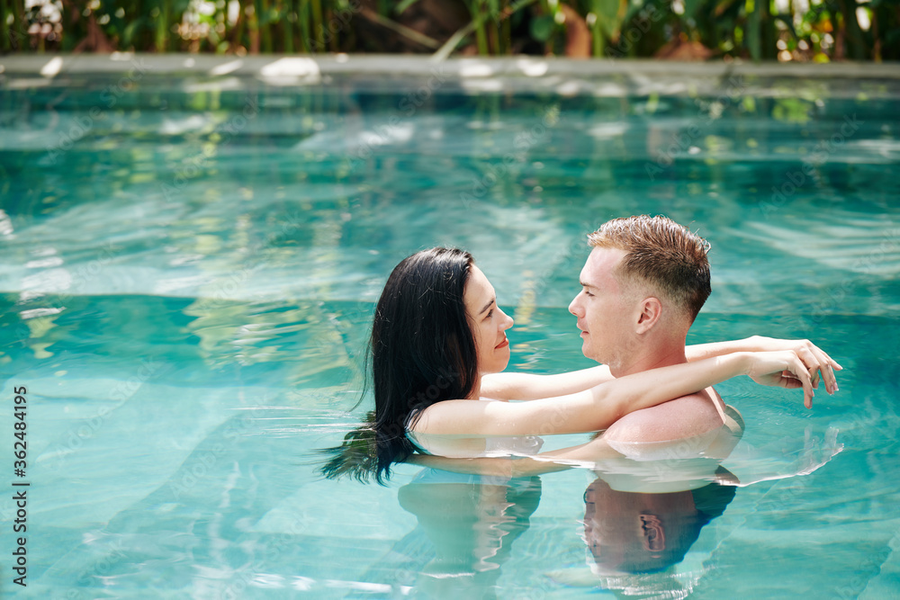 Young couple in love standing in swimming pool, hugging and looking at each other