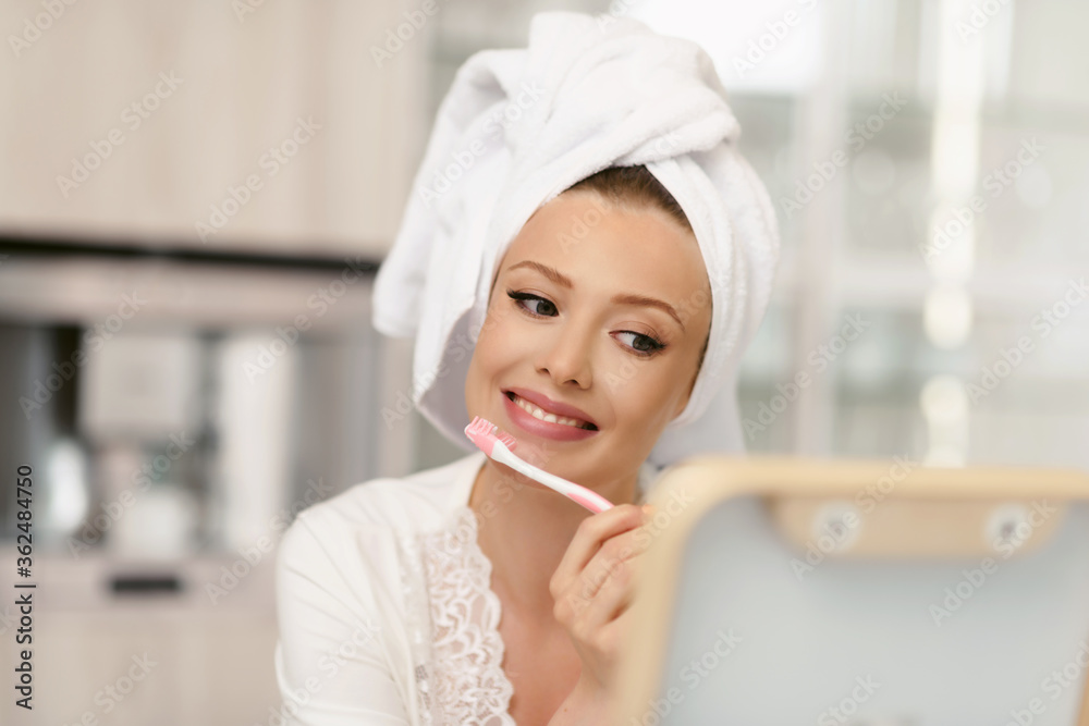 Young attractive woman with a bath towel on her head brushes her teeth in front of a mirror after a shower. Morning oral hygiene at home. Young attractive woman with a bath towel on her head brushes her teeth in front of a mirror after a shower. Morning oral hygiene at home.