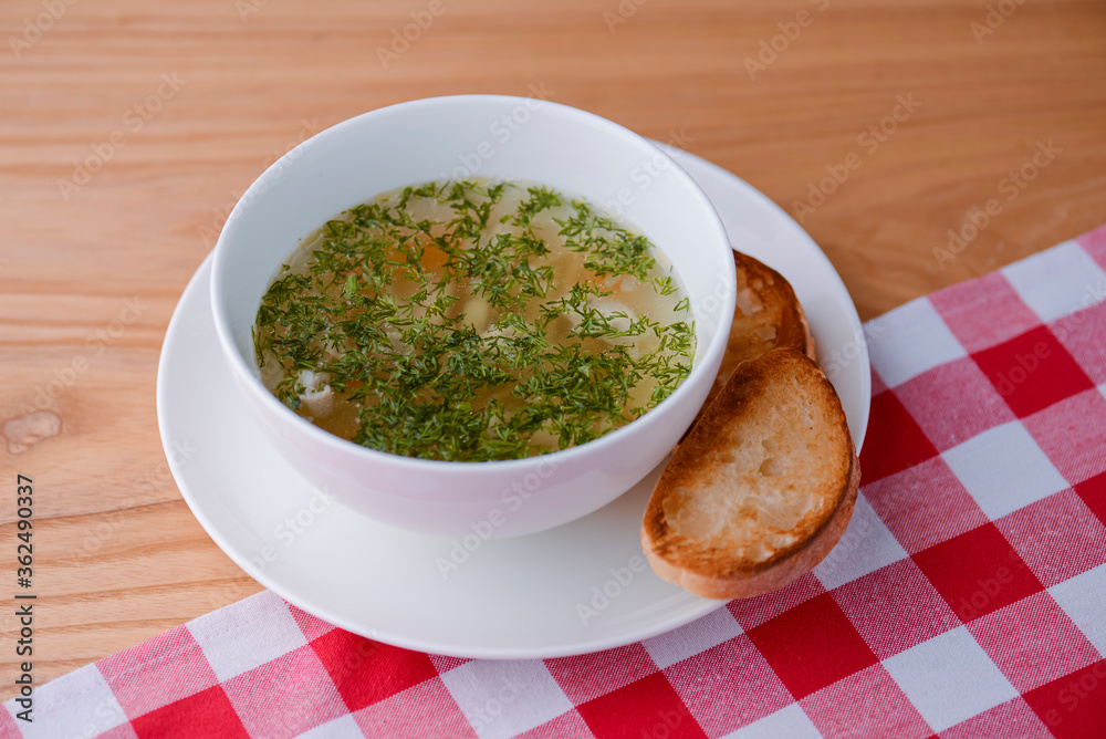 Chicken soup with vegetables and meat served in a white bowl over rustic wooden background with red plaid tablecloth. Chicken soup with vegetables and meat served in a white bowl over rustic wooden background with red plaid tablecloth.