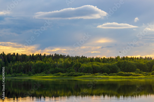 Beautiful evening landscape with a forest lake. Picturesque view of trees on the lake shore and clouds in the sky.