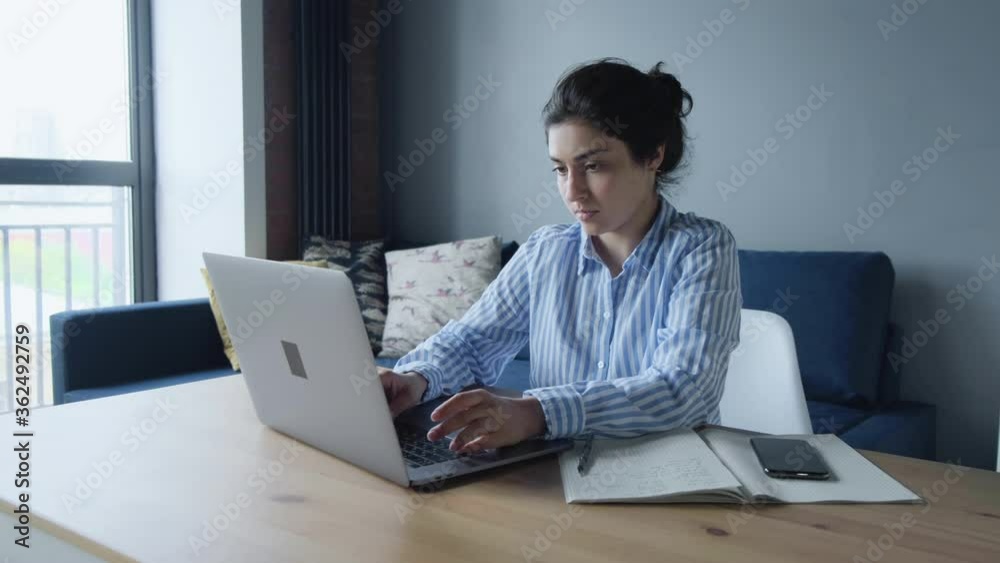 Serious Indian Woman Working Online On A Computer Sitting At A Table In The Kitchen Of The House Next To A Coffee Mug Serious Indian Woman Working Online On A Computer Sitting At A Table In The Kitchen Of The House Next To A Coffee Mug