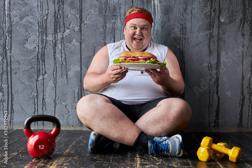 obese young caucasian man encroaches on a big sandwich, fat man can not adhere to a healthy lifestyle and diet