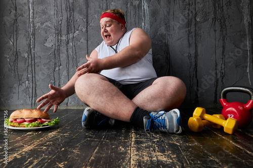 fat caucasian man cannot control himself, hungry man has a mad desire to eat sandwich. male in sportive wear sits on the floor, isolated