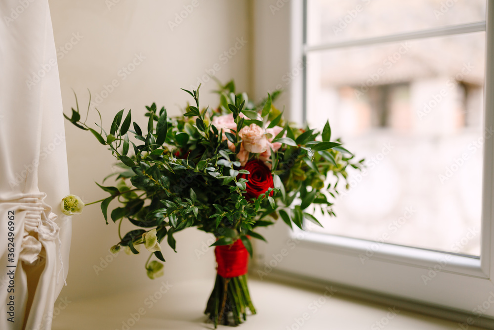 bridal bouquet of red and pink roses, boxwood branches, not blooming buds of white flowers and red ribbons on the window