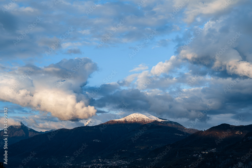 Snow-covered peak of the mountain is illuminated by the sun, the velvet white clouds on the blue sky, on Lake Como in Italy.