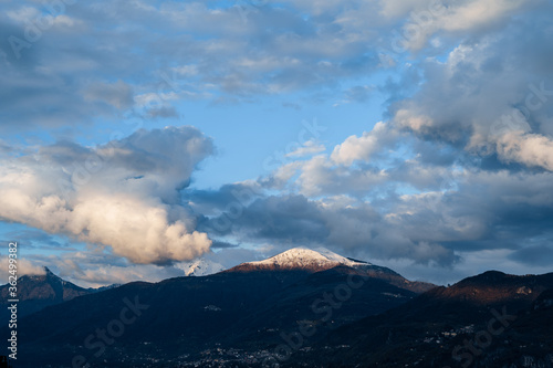 Snow-covered peak of the mountain is illuminated by the sun, the velvet white clouds on the blue sky, on Lake Como in Italy.