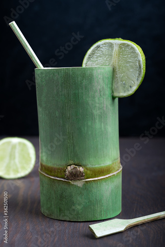 Single green bamboo glass with lime slices and bamboo spoons is standing on dark wooden table at kitchen photo