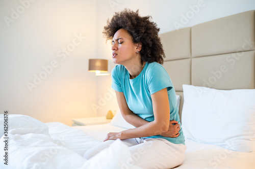 Young woman suffering from abdominal pain while sitting on bed at home. Woman sitting on bed and having stomach ache. Young woman suffering from abdominal pain photo