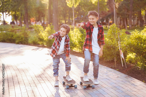 Two pretty brothers of Caucasian nationality standing in the sunny summer park on skateboards. Dressed in jeans and plaid red shirts with white T-shirts. Ponting fingers to camera and laughing