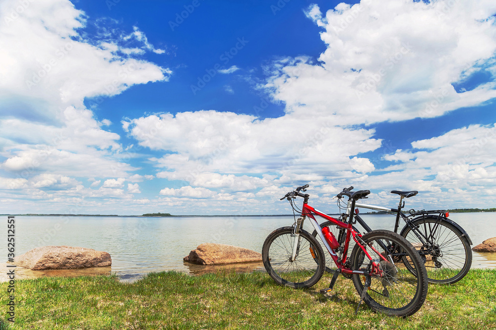 Fototapeta premium Two bikes are on the picturesque background with blue sky, white clouds, water and green grass.