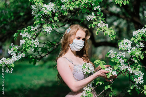 Beautiful young bride in a wedding dress and a white medical mask on her face near a blooming apple. © Ivan Zelenin