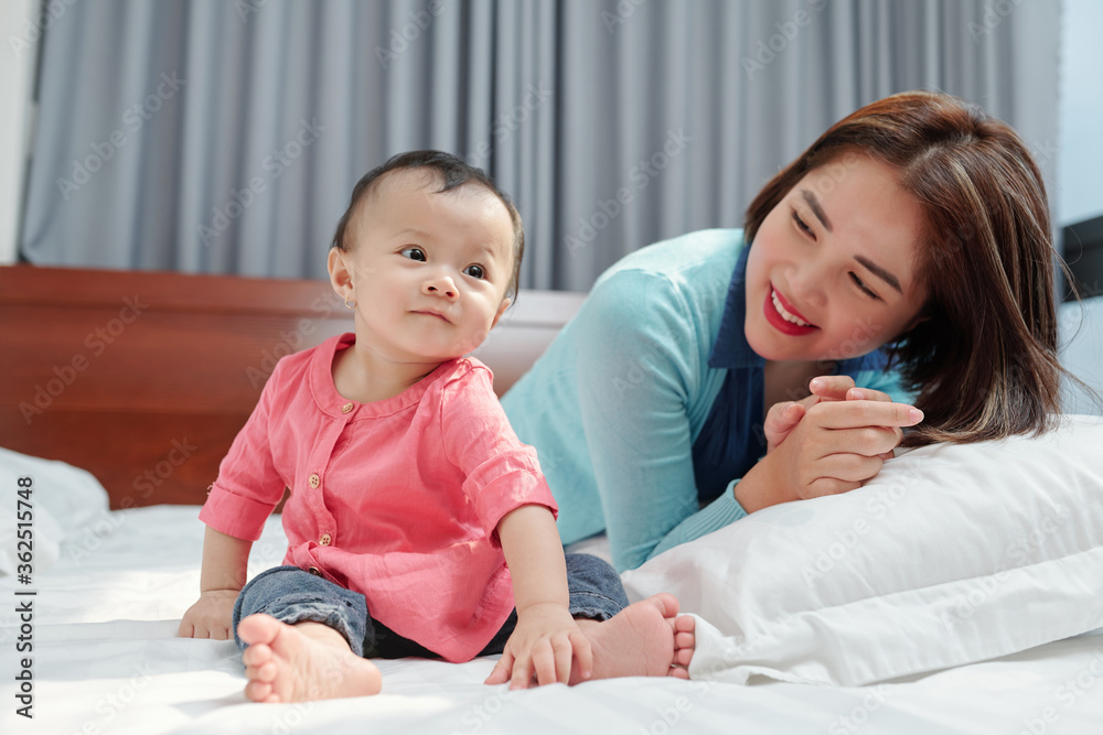 Happy pretty young Vietnamese woman looking at her smiling little daughter sitting on bed Happy pretty young Vietnamese woman looking at her smiling little daughter sitting on bed