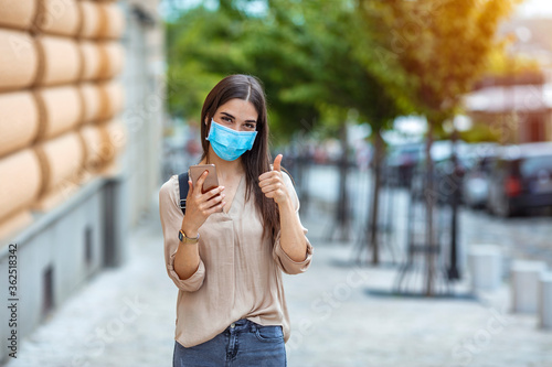 Woman wearing face mask protect from virus showing thumb up. Woman wearing medical face protection mask using smartphone and showing ok sign. photo