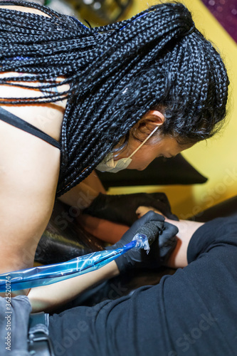 Tattoo artist on action, doing a tattoo on customer's arm, artist is recognizable, wearing a face mask