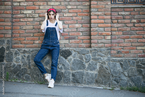 Slim sexy brunette girl wearing woman's denim overall over green tree wall texture in urban area