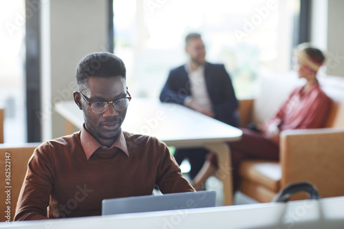 Wallpaper Mural Portrait of young African-American man dressed in business casual using laptop in office or coworking space with people in background, copy space Torontodigital.ca