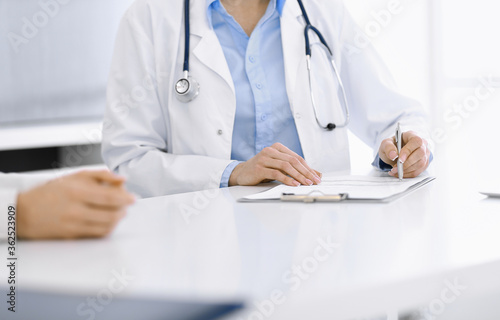 Unknown woman-doctor and female patient sitting and talking at medical examination in clinic, close-up. Therapist wearing blue blouse is filling up medication history record. Medicine concept