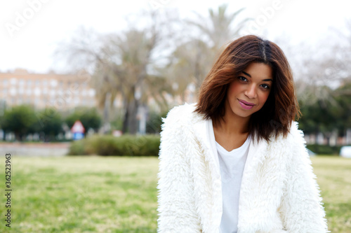 Charming female hipster in stylish clothing posing to the camera while sitting outdoors in beautiful park, afro american woman with brunette hair with copy space for your text message by side