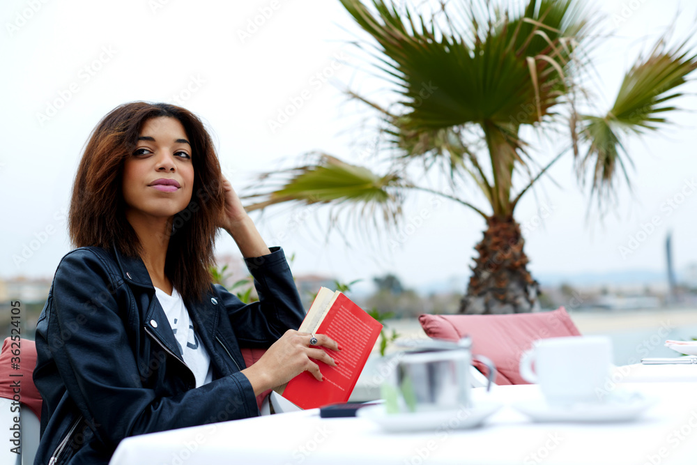 Carefree young woman enjoying a good day with book while sitting at sidewalk cafe near the beach, attractive afro american woman looking for someone in restaurant Carefree young woman enjoying a good day with book while sitting at sidewalk cafe near the beach, attractive afro american woman looking for someone in restaurant