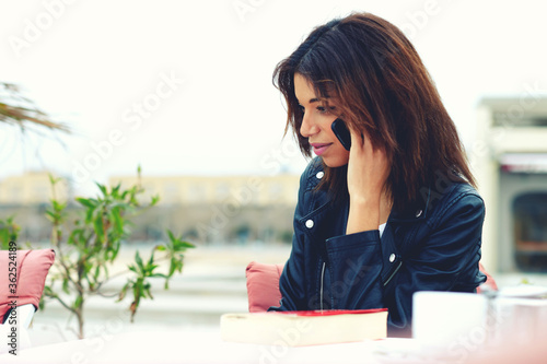 Attractive afro american woman talking on the smart phone while sitting at coffee shop terrace, young female having cell phone conversation sitting at restaurant table during her recreation weekend photo