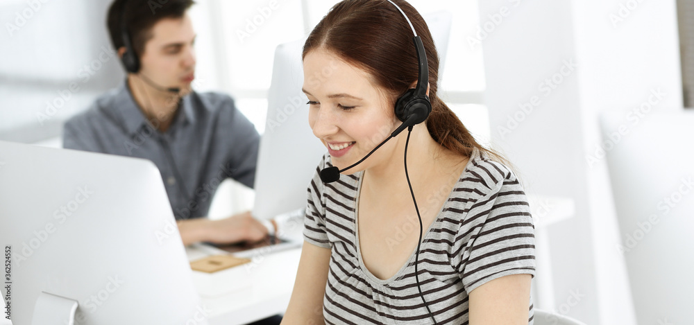 Casual dressed young woman using headset and computer while talking with customers online. Group of operators at work. Call center, business concept Casual dressed young woman using headset and computer while talking with customers online. Group of operators at work. Call center, business concept