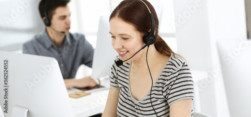 Casual dressed young woman using headset and computer while talking with customers online. Group of operators at work. Call center, business concept