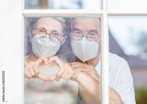 Elderly couple wearing protective face masks watch through their home window and show heart sign during the coronavirus epidemic
