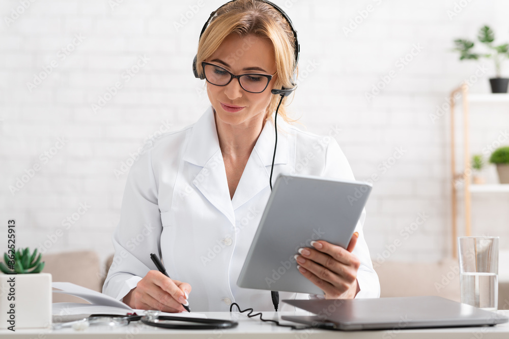 Female doctor makes notes in notebook, holds tablet in hand in interior of office Female doctor makes notes in notebook, holds tablet in hand in interior of office