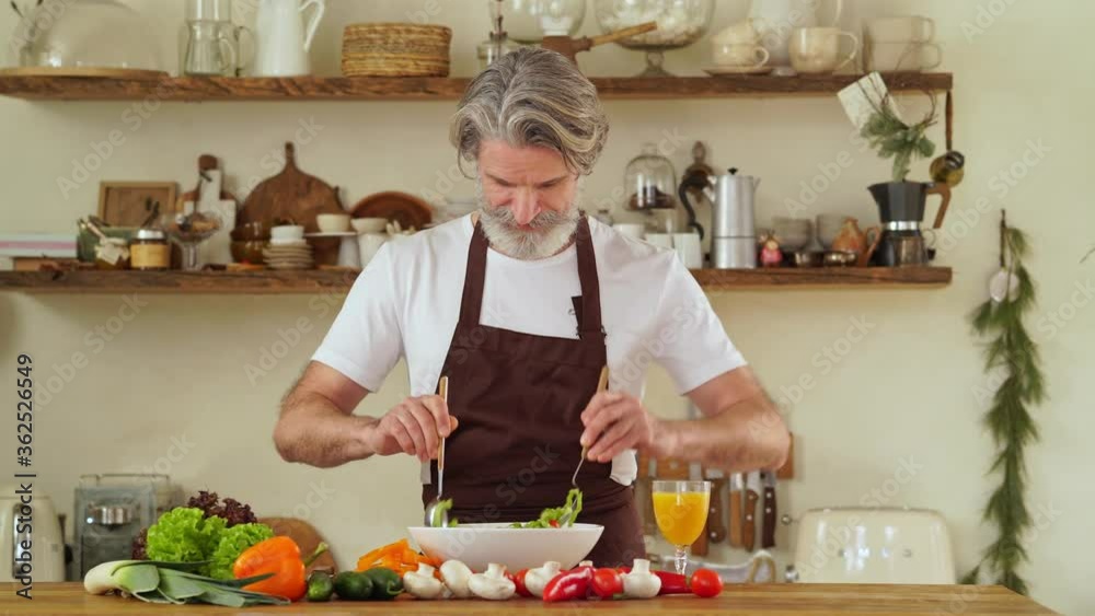 A happy mature man in apron is mixing salad at a cozy kitchen A happy mature man in apron is mixing salad at a cozy kitchen
