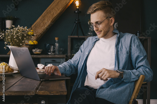 A young guy is sitting at the kitchen table and working on a laptop! A handsome young businessman works from home in a blue shirt