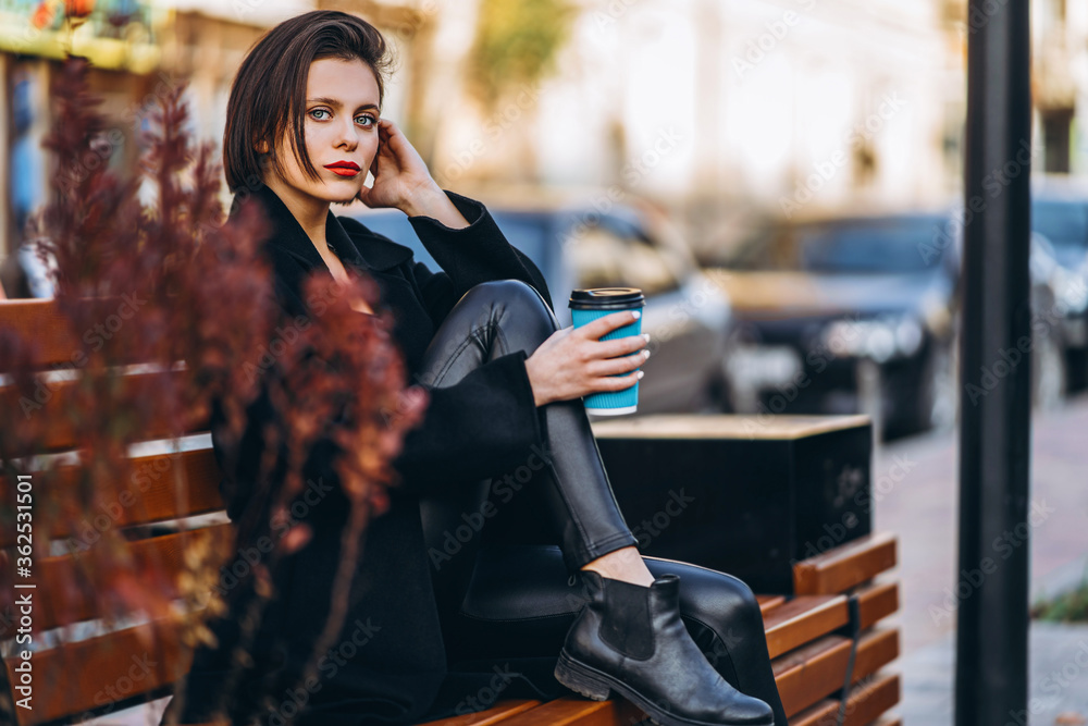 Fototapeta premium Young woman with short haircut, dressed in black, sits on a bench in the city. A woman holds a cup of coffee and looks into the camera