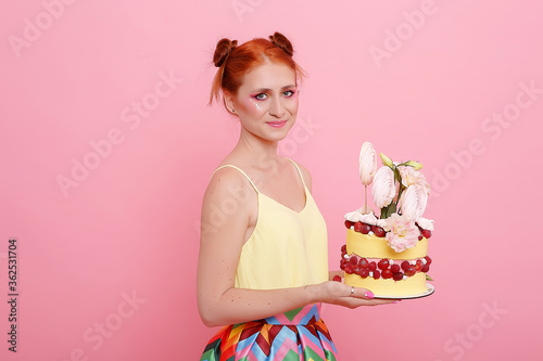 Red-haired girl in colorful dress on a pink background with sweet cake.