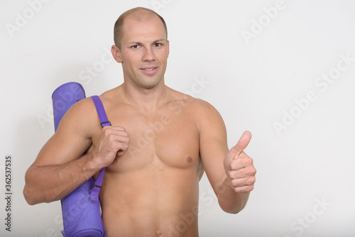 Happy young muscular bald man carrying yoga mat and giving thumbs up shirtless
