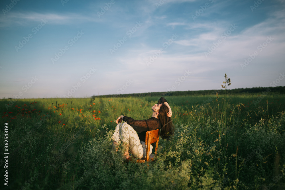 Slow living. Stylish elegant girl sitting on rustic chair in summer meadow with flowers. Fashionable young woman relaxing in field in evening light. Creative beautiful image.