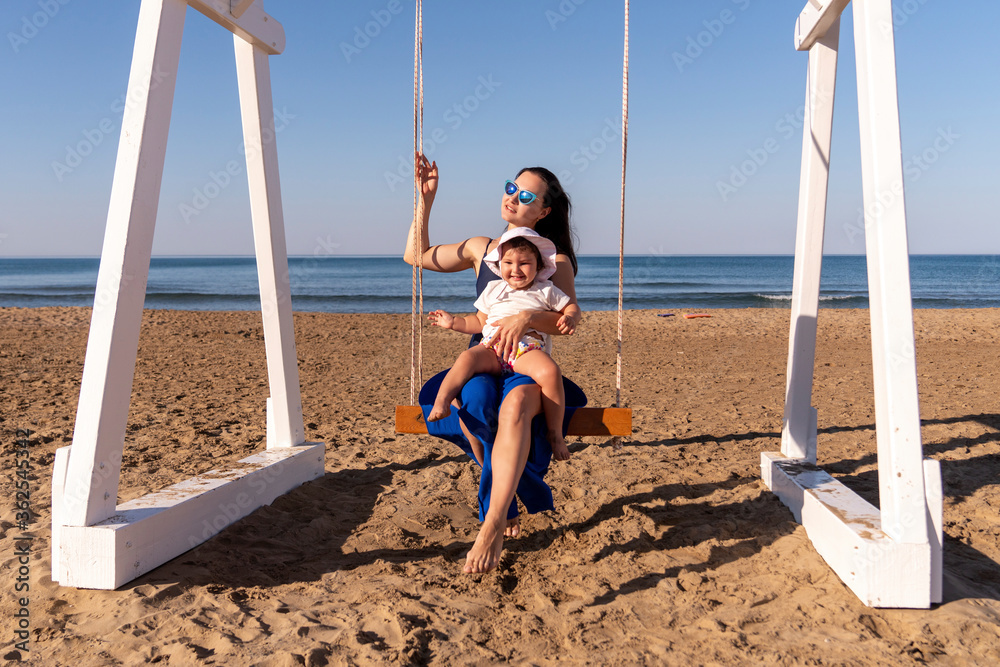 Fototapeta premium Beautiful woman swinging with her baby at the fine sandy Beach enjoying the idyllic scene of the sun and sea. Beautyful sandy beach in Antalya.