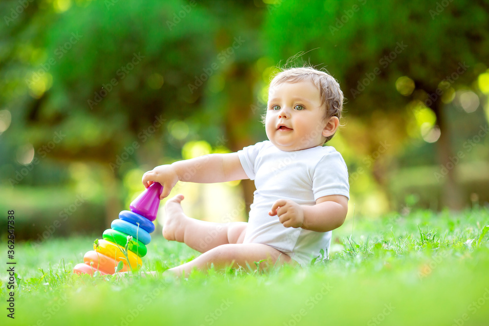 Cute toddler boy in white bodysuit sitting on the green grass in summer plays with a dress multi-colored pyramid. The child plays with the developing toy. Breaking up a child up to a year Cute toddler boy in white bodysuit sitting on the green grass in summer plays with a dress multi-colored pyramid. The child plays with the developing toy. Breaking up a child up to a year