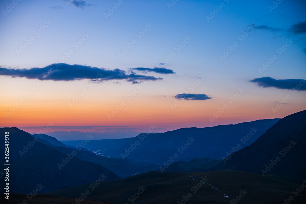 Atmospheric dawn landscape with beautiful mountain river far away in valley among dark mountain silhouettes and vivid orange pink gradient sky with blue clouds of sunset. Dirt road on pass in sunrise.