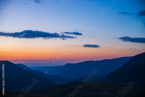 Atmospheric dawn landscape with beautiful mountain river far away in valley among dark mountain silhouettes and vivid orange pink gradient sky with blue clouds of sunset. Dirt road on pass in sunrise.