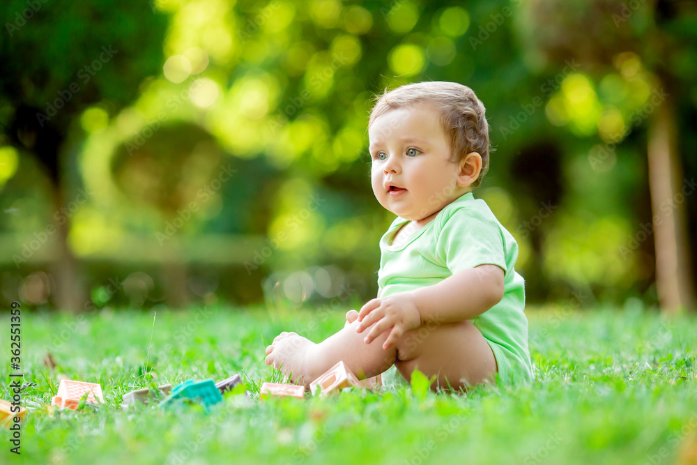 Cute kid in a green bodysuit sitting on the grass in summer plays with developing toys. The baby gnaws silicone cubes. Child development up to one year Cute kid in a green bodysuit sitting on the grass in summer plays with developing toys. The baby gnaws silicone cubes. Child development up to one year