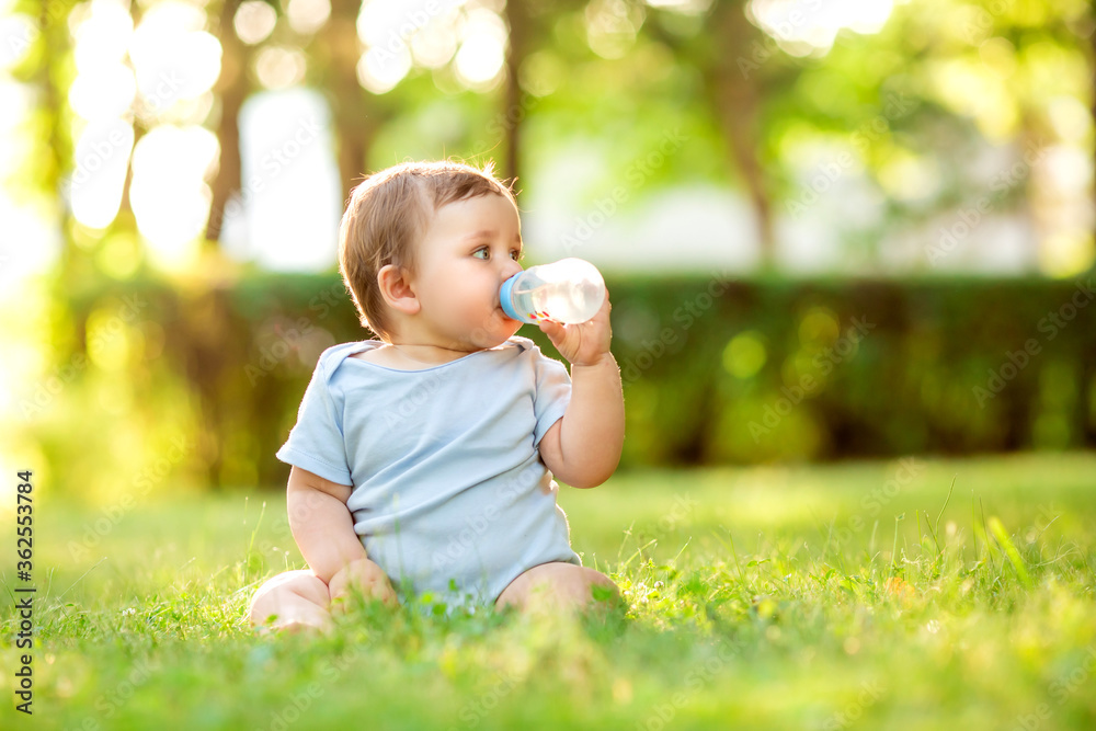 A cute toddler boy in a blue bodysuit sitting on the grass in the summer drinking water from a bottle. Healthy toddler 10 months sitting on the grass drinking water from a bottle A cute toddler boy in a blue bodysuit sitting on the grass in the summer drinking water from a bottle. Healthy toddler 10 months sitting on the grass drinking water from a bottle