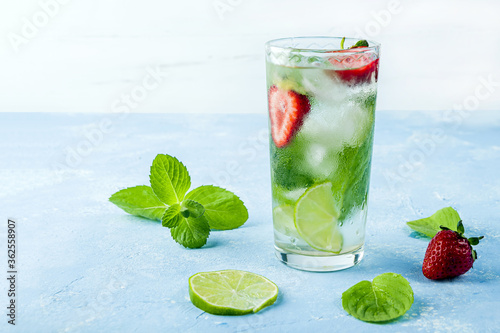 Mojito cocktail with ice cubes. Glass of Summer lemonade or ice tea. Refreshing cool detox drink with strawberry, lime and mint on blue background. Healthy eating. Сopy space for text