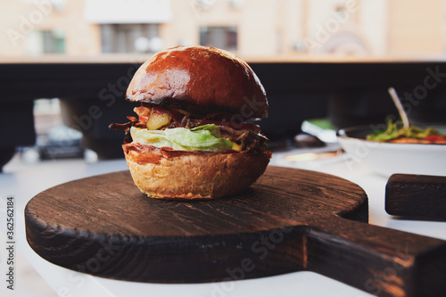 Burger with duck meat on the wooden board at the restaurant. Fresh bun with lettuce and pickled cucumber.