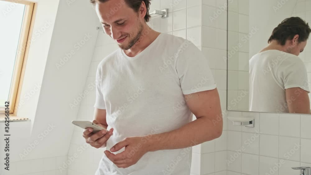 Cheerful handsome man using smartphone while standing in bathroom