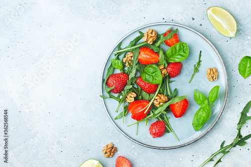 Summer strawberry salad. Fresh arugula, basil, sliced strawberries, nuts with lime on grey stone background. Healthy vegan food concept. Flat lay, top view, copy space