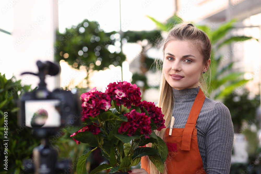 Happy pretty woman in red apron holding plants while working in glasshouse and posing at camera