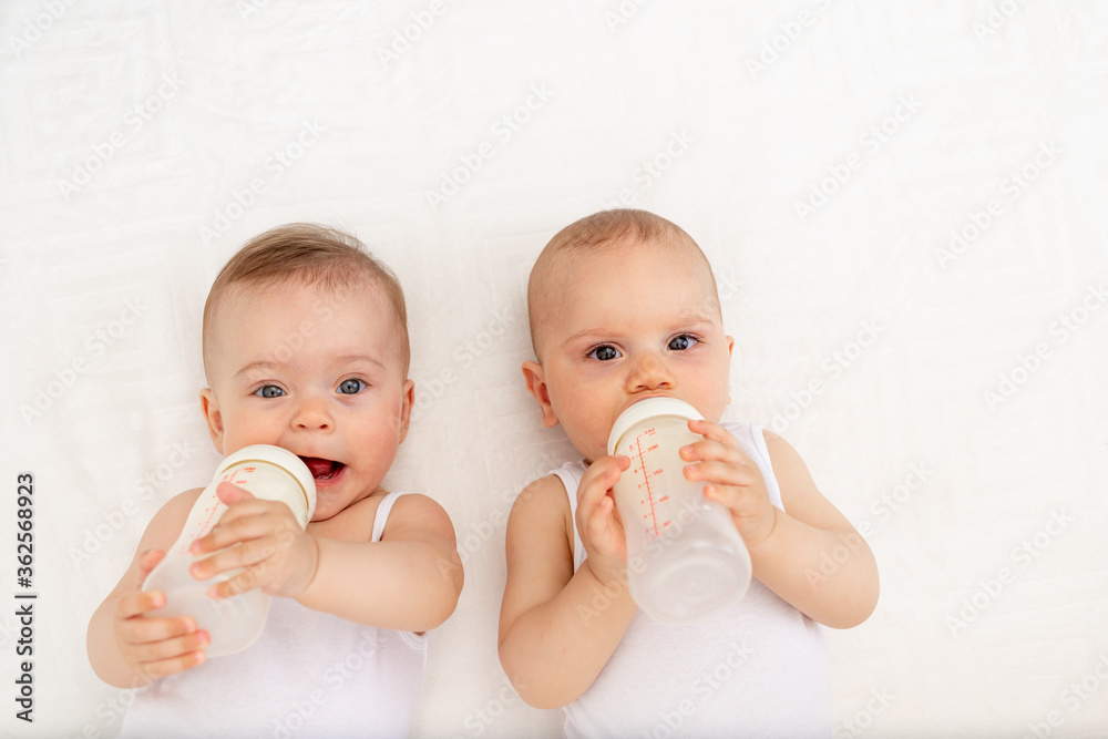 two children a boy and a girl-twins of 8 months drink milk from a bottle on the bed in the nursery, feeding the baby, baby food concept, top view, place for text two children a boy and a girl-twins of 8 months drink milk from a bottle on the bed in the nursery, feeding the baby, baby food concept, top view, place for text