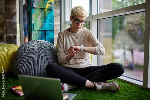 Young hipster girl with stylish short hairstyle checking notification on smartwatch synchronizing data with laptop,female student checking time while working on hometask project in coworking space. photo