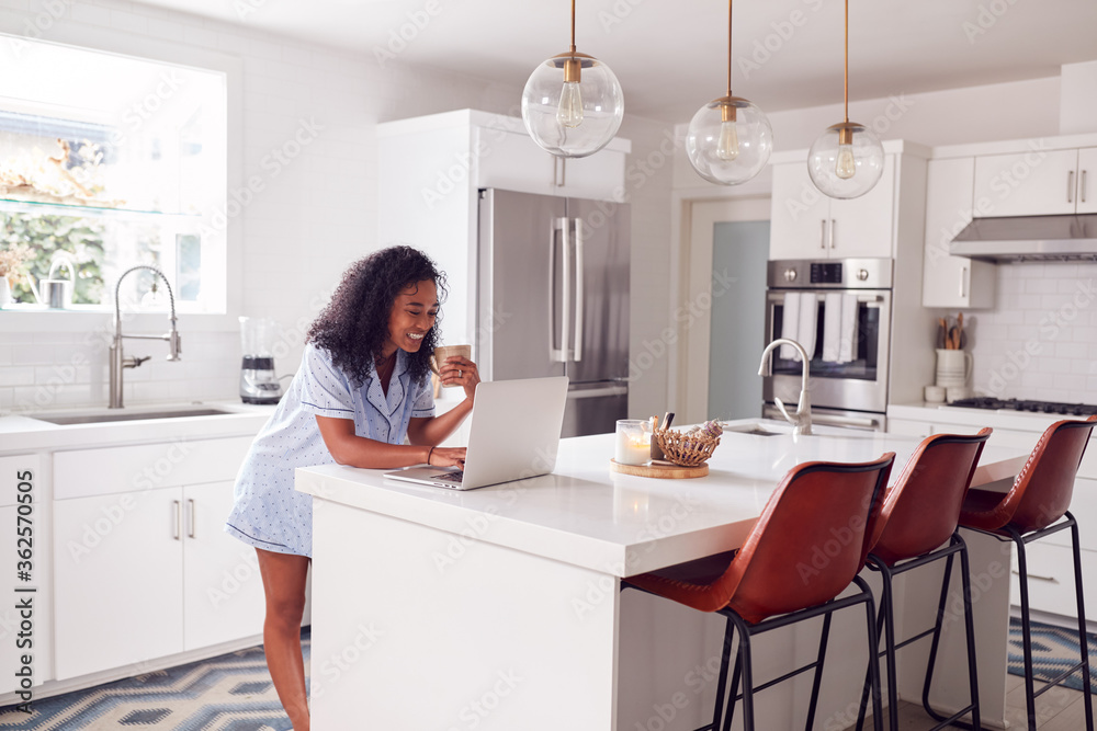 Woman Wearing Pyjamas Standing In Kitchen Working From Home On Laptop Woman Wearing Pyjamas Standing In Kitchen Working From Home On Laptop