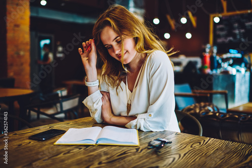 Positive female owner of cafe checking accountings in notepad satisfied with income numbers, smiling beautiful woman pondering on solution while creating business plan for startup looking at notepad